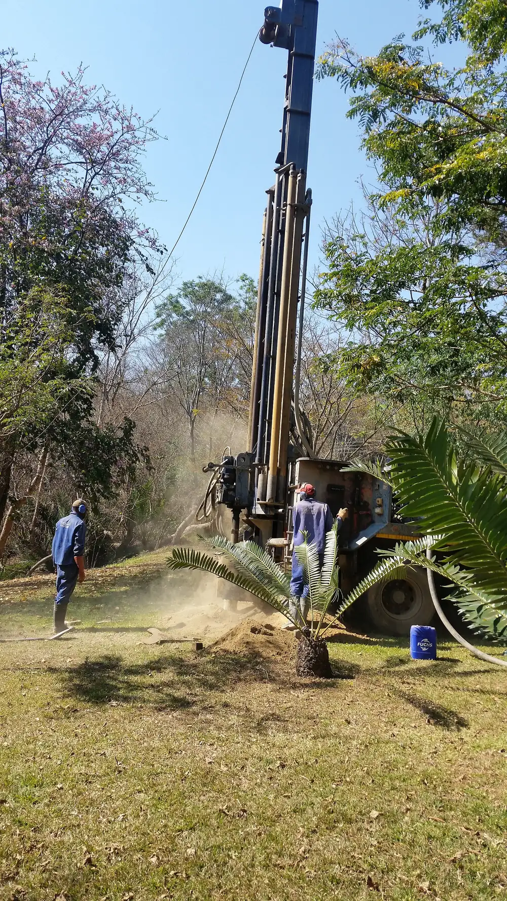 A drilling rig drilling a borehole in a garden, with dust blowing from the hole